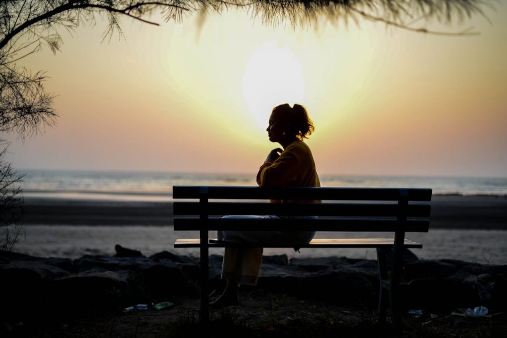 A woman sits alone on a bench by the sea with a sunset in the background